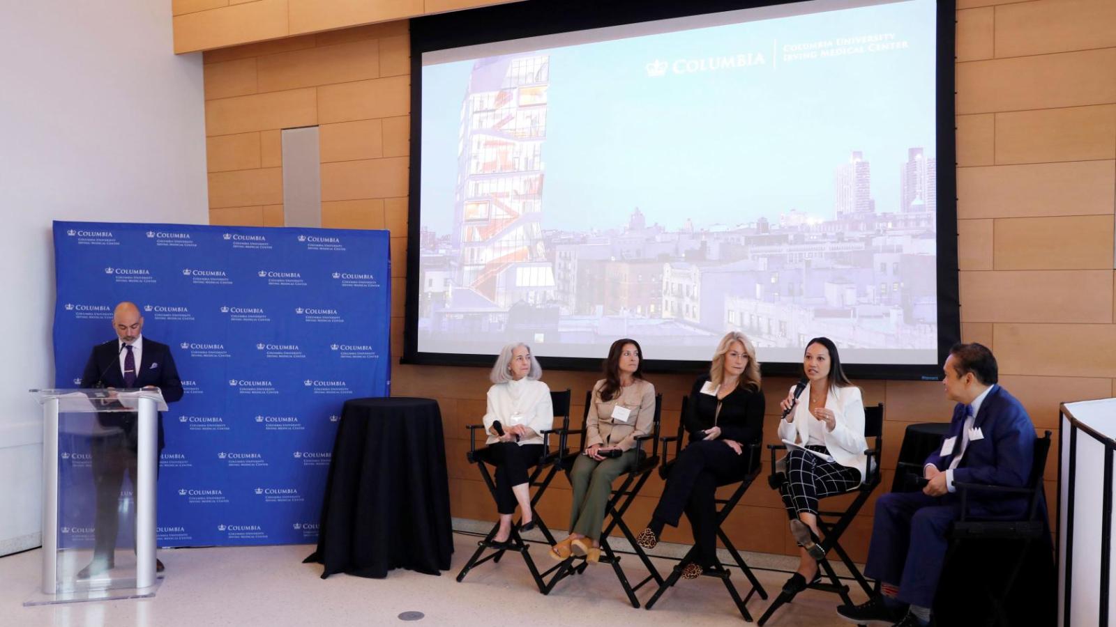 Moderator standing on the left with 5 seated panelists on the right sitting at the front of a conference room