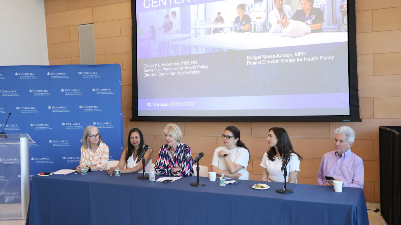A panel of six faculty members and researchers sit at a blue-draped table in front of a large screen