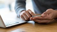 Close-up of unrecognizable man in grey sweater sitting at wooden table next to laptop and holding cell phone in both hands