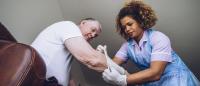 Nurse in gown is helping a man sitting in bed put on a bandage at home.